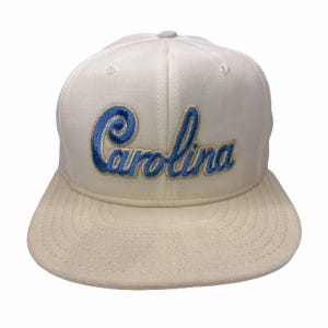K&ouml;nnte beinhalten: Cremefarbene Baseballkappe mit flachem Schirm. Das Wort "Carolina" ist in blauer Schrift auf die Vorderseite gestickt. Die Kappe hat eine strukturierte Krone und einen grauen Knopf oben.