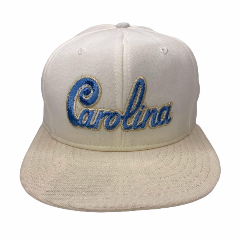 K&ouml;nnte beinhalten: Cremefarbene Baseballkappe mit flachem Schirm. Das Wort "Carolina" ist in blauer Schrift auf die Vorderseite gestickt. Die Kappe hat eine strukturierte Krone und einen grauen Knopf oben.
