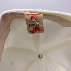 K&ouml;nnte beinhalten: Nahaufnahme der Innenseite einer cremefarbenen Baseballkappe. Ein braunes Etikett mit der Aufschrift "MADE IN AMERICAN WORKERS 100% NYLON DOUBLE KNIT REGULAR" mit einem rot-wei&szlig;en Logo. Die Kappe ist aus Nylon.