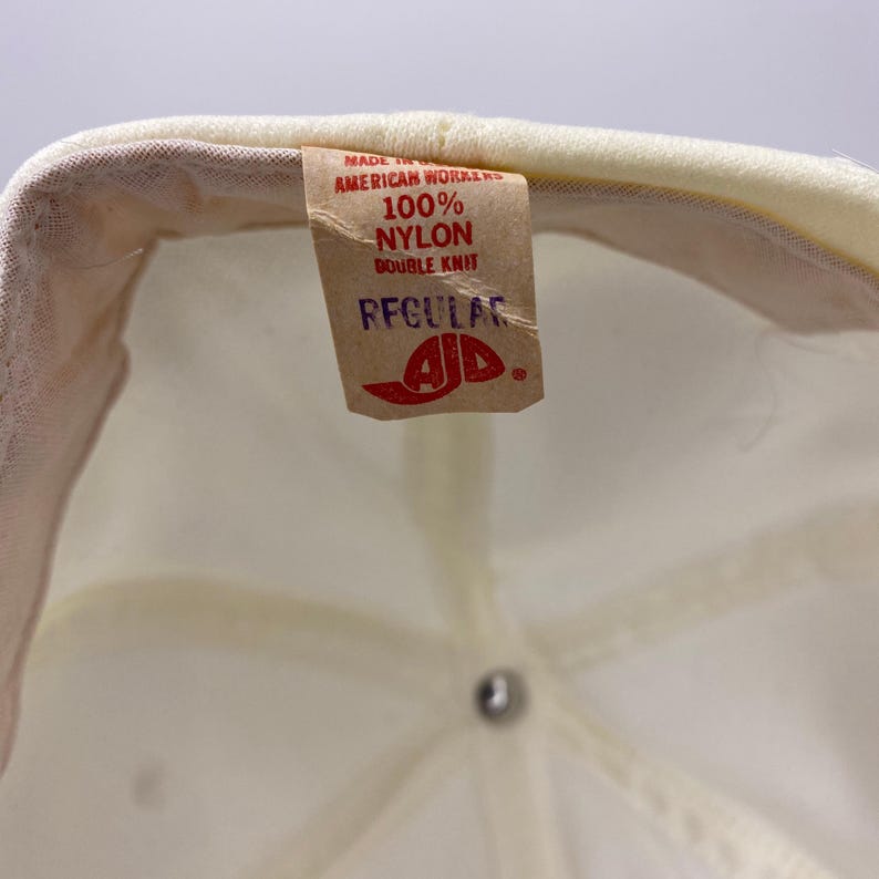 K&ouml;nnte beinhalten: Nahaufnahme der Innenseite einer cremefarbenen Baseballkappe. Ein braunes Etikett mit der Aufschrift "MADE IN AMERICAN WORKERS 100% NYLON DOUBLE KNIT REGULAR" mit einem rot-wei&szlig;en Logo. Die Kappe ist aus Nylon.