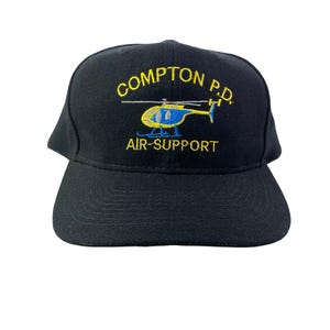 May include: Black baseball cap with the text "COMPTON P.D." in yellow, and a blue and yellow helicopter graphic. The words "AIR-SUPPORT" are embroidered below the helicopter.