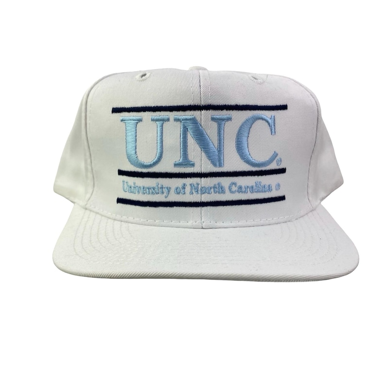 Puede incluir: Gorra de b&eacute;isbol blanca con visera plana. La gorra presenta las letras bordadas en azul claro "UNC" sobre las palabras "University of North Carolina" en fuente azul oscuro. La gorra tiene una corona estructurada y cierre trasero a presi&oacute;n.