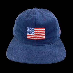 May include: A navy blue baseball cap with a curved brim. The cap features an embroidered American flag patch in red, white, and blue on the front. The hat is made of a soft fabric and has a classic, casual style.