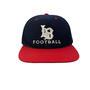 May include: A navy blue baseball cap with a red brim. The cap features a white logo and the word "FOOTBALL" in white block letters. The cap has a red button on top.
