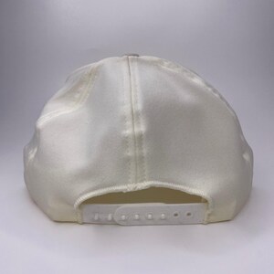 K&ouml;nnte beinhalten: Eine cremefarbene Baseballkappe mit gebogenem Schirm und verstellbarem Schnappverschluss. Die Kappe besteht aus einem weichen, strukturierten Stoff und hat ein klassisches Sechs-Panel-Design. Auf der R&uuml;ckseite befindet sich ein wei&szlig;er Kunststoffriemen zur Gr&ouml;&szlig;enanpassung.