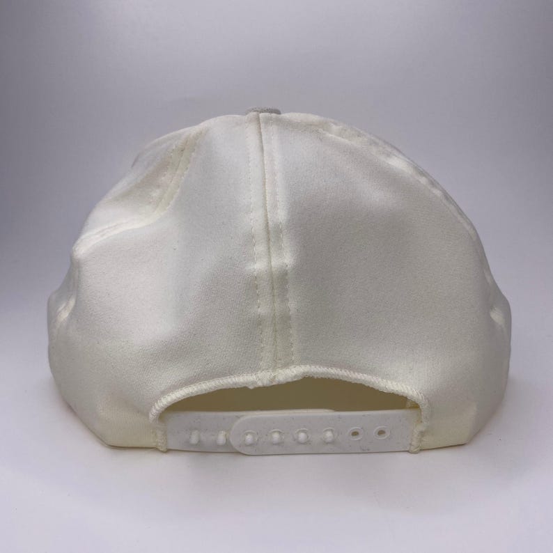 K&ouml;nnte beinhalten: Eine cremefarbene Baseballkappe mit gebogenem Schirm und verstellbarem Schnappverschluss. Die Kappe besteht aus einem weichen, strukturierten Stoff und hat ein klassisches Sechs-Panel-Design. Auf der R&uuml;ckseite befindet sich ein wei&szlig;er Kunststoffriemen zur Gr&ouml;&szlig;enanpassung.