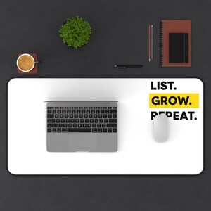 Puede incluir: Una alfombrilla de escritorio blanca con las palabras "LIST. GROW. REPEAT." en negro y amarillo. Un ordenador portátil plateado, un ratón blanco y otros artículos de oficina están sobre una superficie negra.