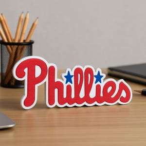 May include: A red and white Phillies logo, with two blue stars, is displayed on a wooden desk. The word "Phillies" is in a bold, stylized font. Pencils in a black holder are in the background.