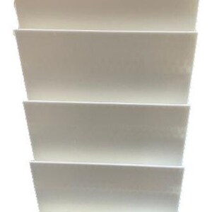 May include: A stack of four off-white, rectangular, tiered shelves. Each shelf is slightly angled, creating a stepped appearance. The shelves are made of a solid material and are suitable for displaying items.