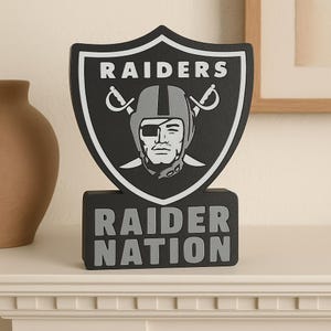 Puede incluir: Un logotipo en blanco y negro del equipo Raiders que se muestra en una placa en forma de escudo. La placa presenta el nombre del equipo y las palabras "RAIDER NATION". El logotipo incluye una cara con casco, parche en el ojo y espadas cruzadas.