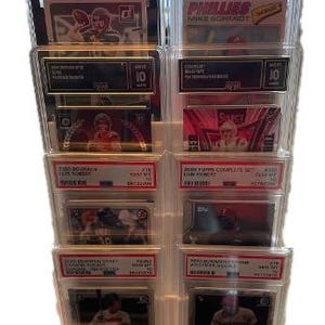 May include: A clear plastic display stand holding multiple graded sports trading cards. The cards feature various players and teams, with visible grading labels and text. The stand is beige and has multiple tiers.