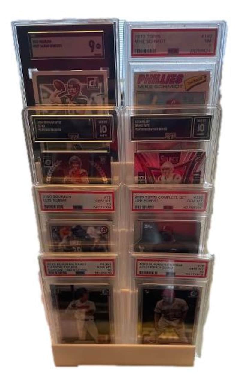 May include: A clear plastic display stand holding multiple graded sports trading cards. The cards feature various players and teams, with visible grading labels and text. The stand is beige and has multiple tiers.