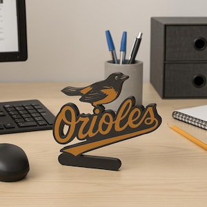 May include: A black and orange Orioles logo with a bird perched on top. The word "Orioles" is in orange script with a black outline. The logo is on a wooden base, and is displayed on a desk.