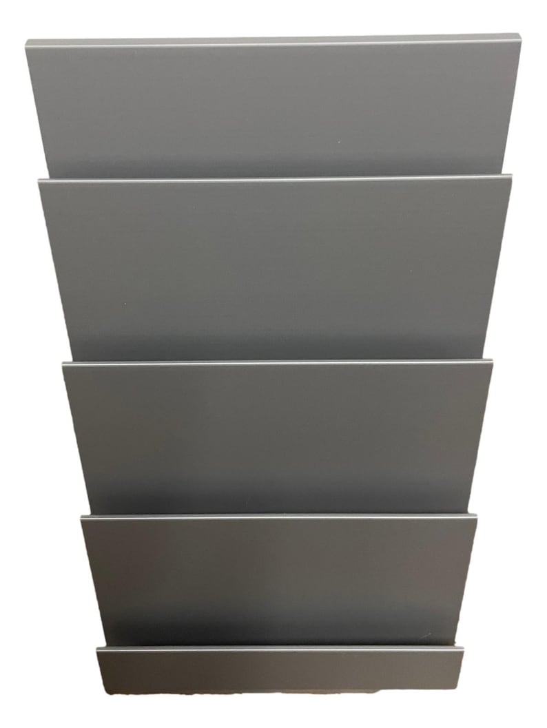 May include: A gray metal wall-mounted magazine rack with five horizontal shelves. The shelves are angled slightly upward, creating a tiered design. The rack is made of a smooth, solid material and is designed to hold magazines or other reading materials.