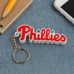 May include: A white and red Phillies keychain with a silver chain and ring. The word "Phillies" is written in a script font with blue star accents. The keychain is on a blue denim surface.