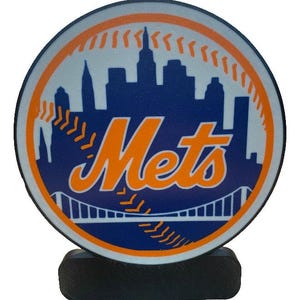 May include: A round Mets logo on a black base. The logo features the word "Mets" in orange script over a blue background with a white skyline and orange baseball stitching.