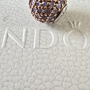 Op de afbeelding: Een ronde bedel met een goudkleurige metalen basis en een oppervlak bedekt met kleine, lichtpaarse stenen. De bedel is geplaatst op een getextureerd wit oppervlak. Dit is een sieraadaccessoire.