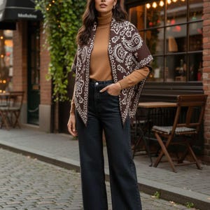 May include: A woman wearing a brown paisley print shawl over a tan turtleneck sweater and dark wash denim jeans. The shawl has a white paisley pattern on a brown background. She is standing on a cobblestone street.