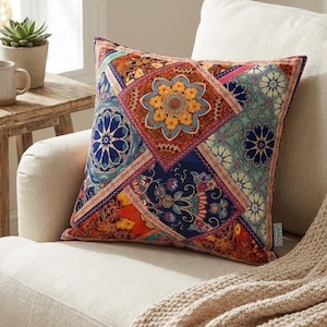 May include: Square decorative pillow with a patchwork design. The pillow features a central orange floral pattern surrounded by blue, teal, and orange geometric shapes. The pillow is on a cream-colored armchair with a beige throw blanket.