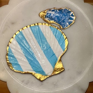 Puede incluir: Dos cuencos decorativos con forma de concha. Uno está pintado con rayas azules y blancas y ribete dorado. El otro es una concha de ostra más pequeña con un diseño floral azul y ribete dorado. Ambos están sobre una superficie blanca.