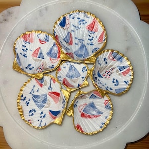Puede incluir: Seis conchas de vieira con temática náutica. Las conchas blancas presentan un patrón de veleros rojos y azules, con bordes dorados. Están dispuestas sobre un plato blanco con bordes festoneados.