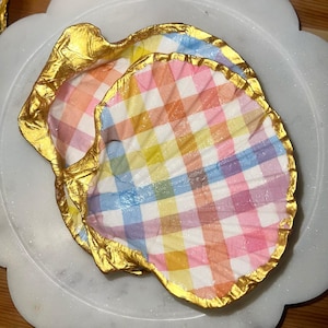 Puede incluir: Dos platos decorativos con forma de concha con un colorido estampado de cuadros en rosa, amarillo, azul y naranja. Los bordes están adornados con pan de oro. Los platos están sobre un plato blanco con borde festoneado.