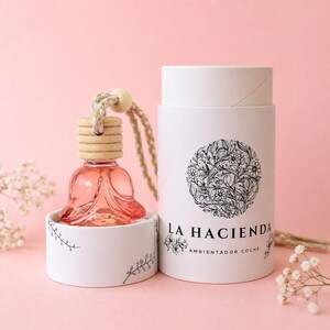 Puede incluir: Un ambientador para coche de cristal rosa con una tapa de madera y una cuerda color crema. El ambientador está en un recipiente blanco con un diseño floral y las palabras "LA HACIENDA". Una caja cilíndrica a juego está al lado.