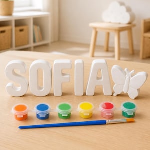 Könnte beinhalten: Ein Set aus weißen, dreidimensionalen Buchstaben, die den Namen SOFIA bilden, ergänzt durch eine schmetterlingsförmige Figur. Unter den Buchstaben befinden sich sechs kleine, verbundene Kunststofftöpfe mit orangefarbener, blauer, grüner, gelber, roter und grüner Farbe sowie ein Pinsel.
