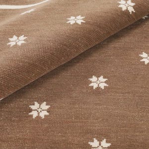 May include: A brown woven rug featuring a repeating pattern of small white four-pointed star or snowflake motifs. The rug has a thick white border along the edge and is folded over at the top corner to reveal the textured fabric surface.