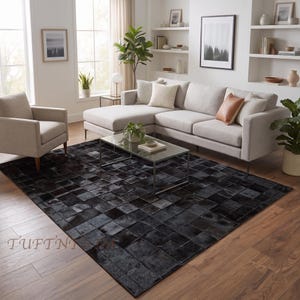 May include: A large rectangular area rug featuring a patchwork design of square black and dark gray leather tiles. The rug has a smooth, low-pile texture and is positioned on a wooden floor beneath a glass coffee table in a living room.