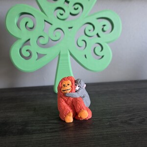 May include: A small, orange orangutan figurine hugging a gray monkey figurine. The figurines are in front of a large, green, wooden shamrock decoration with intricate cut-out designs. The background is a dark surface.
