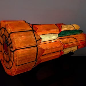 May include: A cylindrical lamp featuring a stained glass-style design with orange, yellow, and green panels separated by thick black lines. The structure has a circular front face with a radial pattern and a textured, multi-faceted exterior surface.
