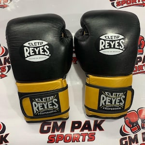 Peut inclure: Paire de gants de boxe noirs et dorés avec le logo "CLETO REYES". Les gants ont une partie supérieure noire et un support de poignet doré. Le texte "HECHO EN MEXICO" est visible sur les gants.