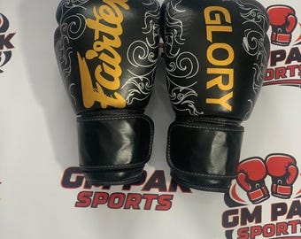 Könnte beinhalten: Paar schwarze Fairtex und Glory Boxhandschuhe. Die Handschuhe haben goldfarbene Schriftzüge und weiße dekorative Wirbel. Die Handschuhe liegen auf einer weißen Oberfläche mit den Worten "GM PAN SPORTS" und einer Grafik von zwei roten Boxhandschuhen.