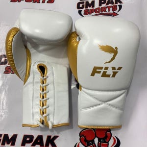 Puede incluir: Un par de guantes de boxeo blancos con ribetes y detalles dorados. Los guantes tienen un cierre con cordones y la palabra "FLY" con una figura alada impresa en la parte delantera. Los guantes están diseñados para deportes y entrenamiento.