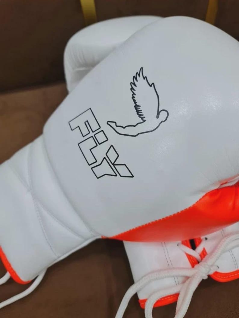May include: White boxing gloves with orange trim and white laces. The gloves have the word "FLY" and a graphic of a bird in black. The gloves are made of a smooth material and are designed for boxing or other combat sports.
