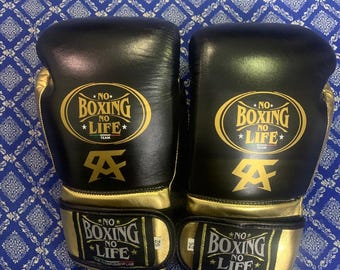 Könnte beinhalten: Ein Paar schwarze und goldene Boxhandschuhe. Die Handschuhe tragen den Text "NO BOXING NO LIFE" in goldener Schrift. Die Handschuhe haben goldene Manschetten und Akzente. Sie liegen auf einem blauen, gemusterten Hintergrund.