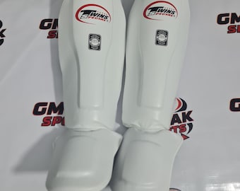 Könnte beinhalten: Paar weiße Schienbeinschützer mit dem Logo "Twins Special" in Rot und Schwarz. Die Schützer sind für Sport und Kampfsport konzipiert, mit Polsterung und einer konturierten Form zum Schutz. Das Logo "GM PAK SPORTS" ist im Hintergrund sichtbar.