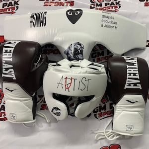 Könnte beinhalten: Boxausrüstung-Set mit einem weißen Kopfschutz mit dem Text "ARTIST", einem weißen Brustschutz mit dem Text "#SWAG" und zwei braunen und weißen Everlast-Boxhandschuhen. Das Set ist für den Sport konzipiert.
