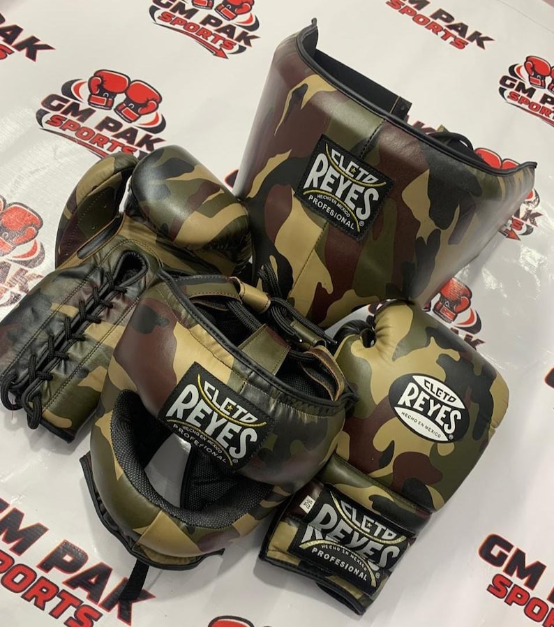 May include: A collection of camouflage boxing gear, including gloves, headgear, and a face protector. The gear features the "CLETO REYES" logo. The color scheme includes shades of green, brown, and tan.