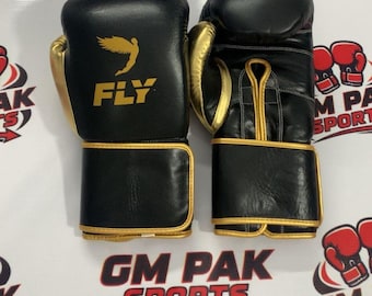 Könnte beinhalten: Ein Paar schwarze Boxhandschuhe mit goldenen Akzenten. Auf den Handschuhen steht das Wort "FLY" und ein geflügeltes Logo. Die Handschuhe werden auf einer weißen Oberfläche mit dem Text "GM PAK SPORTS" in Rot und Weiß präsentiert.