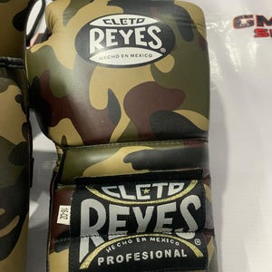 May include: Camouflage-patterned boxing gloves with the "CLETD REYES" logo and "HECHO EN MEXICO" text. The gloves are in shades of green, brown, and tan. The label reads "PROFESIONAL" and "16-OZ".