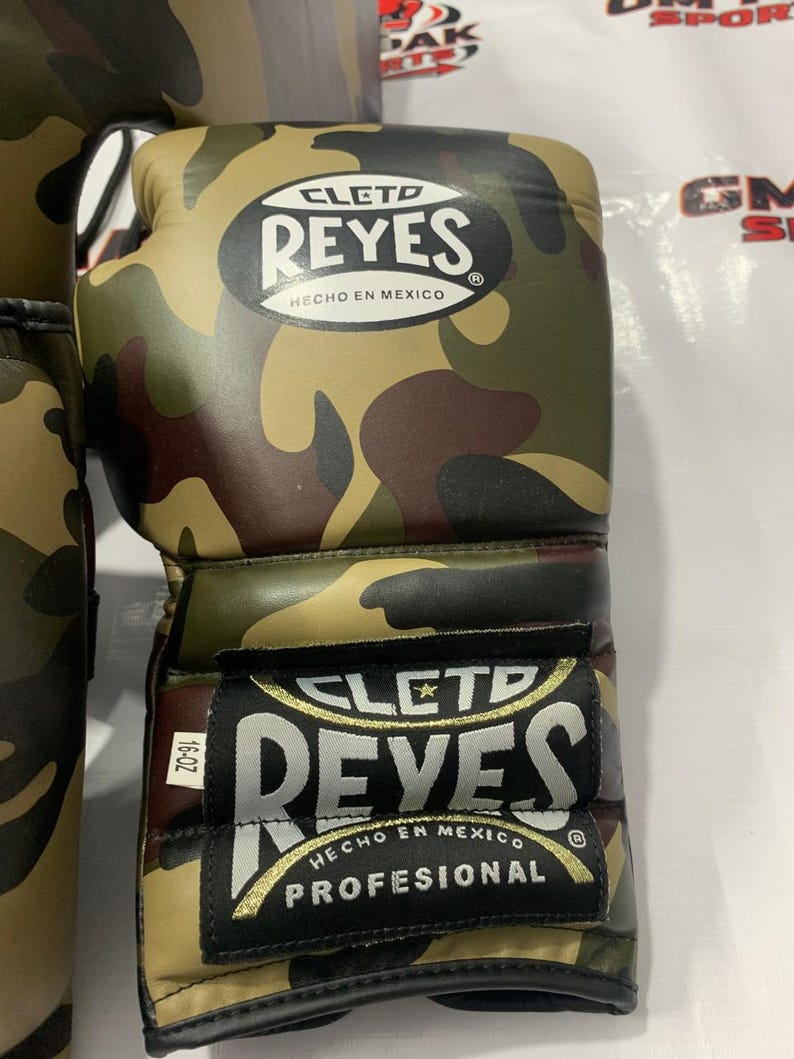 May include: Camouflage-patterned boxing gloves with the "CLETD REYES" logo and "HECHO EN MEXICO" text. The gloves are in shades of green, brown, and tan. The label reads "PROFESIONAL" and "16-OZ".