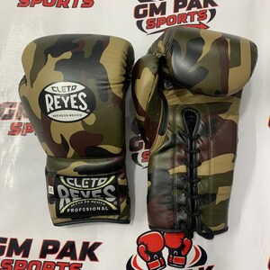 May include: Pair of camouflage boxing gloves with the "CLETO REYES" logo. The gloves are in shades of green, brown, and black. The gloves are laced up and have the text "HECHO EN MEXICO" and "PROFESIONAL". The background has the text "GM PAK SPORTS".
