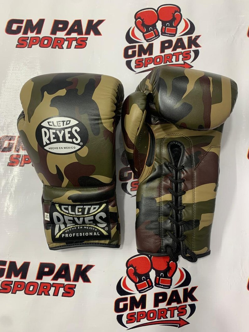 May include: Pair of camouflage boxing gloves with the "CLETO REYES" logo. The gloves are in shades of green, brown, and black. The gloves are laced up and have the text "HECHO EN MEXICO" and "PROFESIONAL". The background has the text "GM PAK SPORTS".