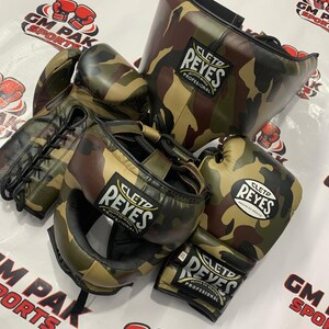 May include: A set of Cleto Reyes boxing gear in a camouflage pattern. The set includes boxing gloves, a head guard, and a face protector. The gloves and headgear feature the Cleto Reyes logo. The background has the text "GM PAK SPORTS".