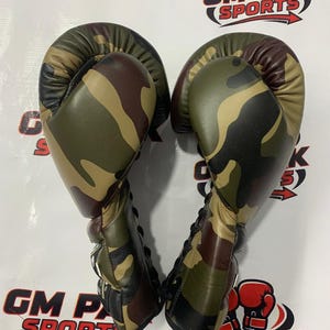 May include: Pair of camouflage boxing gloves. The gloves feature a green, brown, and tan pattern. The text "GM PAK SPORTS" is visible in the background.