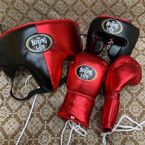 May include: A set of boxing equipment including a red and black headgear, a smaller black headgear, and a pair of metallic red boxing gloves. Each piece features a black and gold oval patch with the text NO BOXING NO LIFE printed in white.