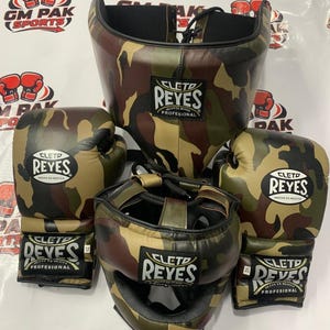 May include: A camouflage boxing set including a head guard and gloves. The gloves and headgear are in a brown, green, and tan camouflage pattern. The brand name "CLETO REYES" is visible on each piece. The set is designed for professional use.