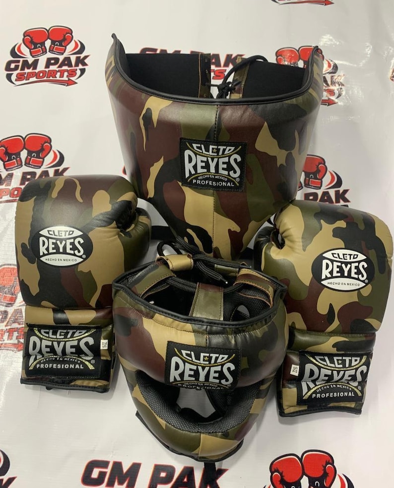 May include: A camouflage boxing set including a head guard and gloves. The gloves and headgear are in a brown, green, and tan camouflage pattern. The brand name "CLETO REYES" is visible on each piece. The set is designed for professional use.